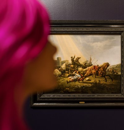 Vrouw met roze haar staat in een museumzaal. Achter haar een schilderij van Cuyp.