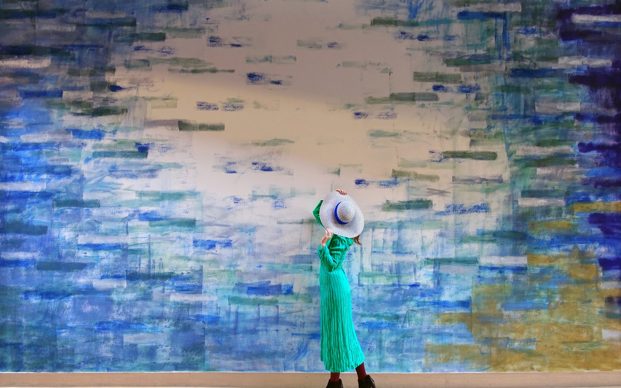 Sofie Kugel loopt in een groene jurk met hoed op langs het blauwe gedeelte van de muurschildering van Robert Zandvliet